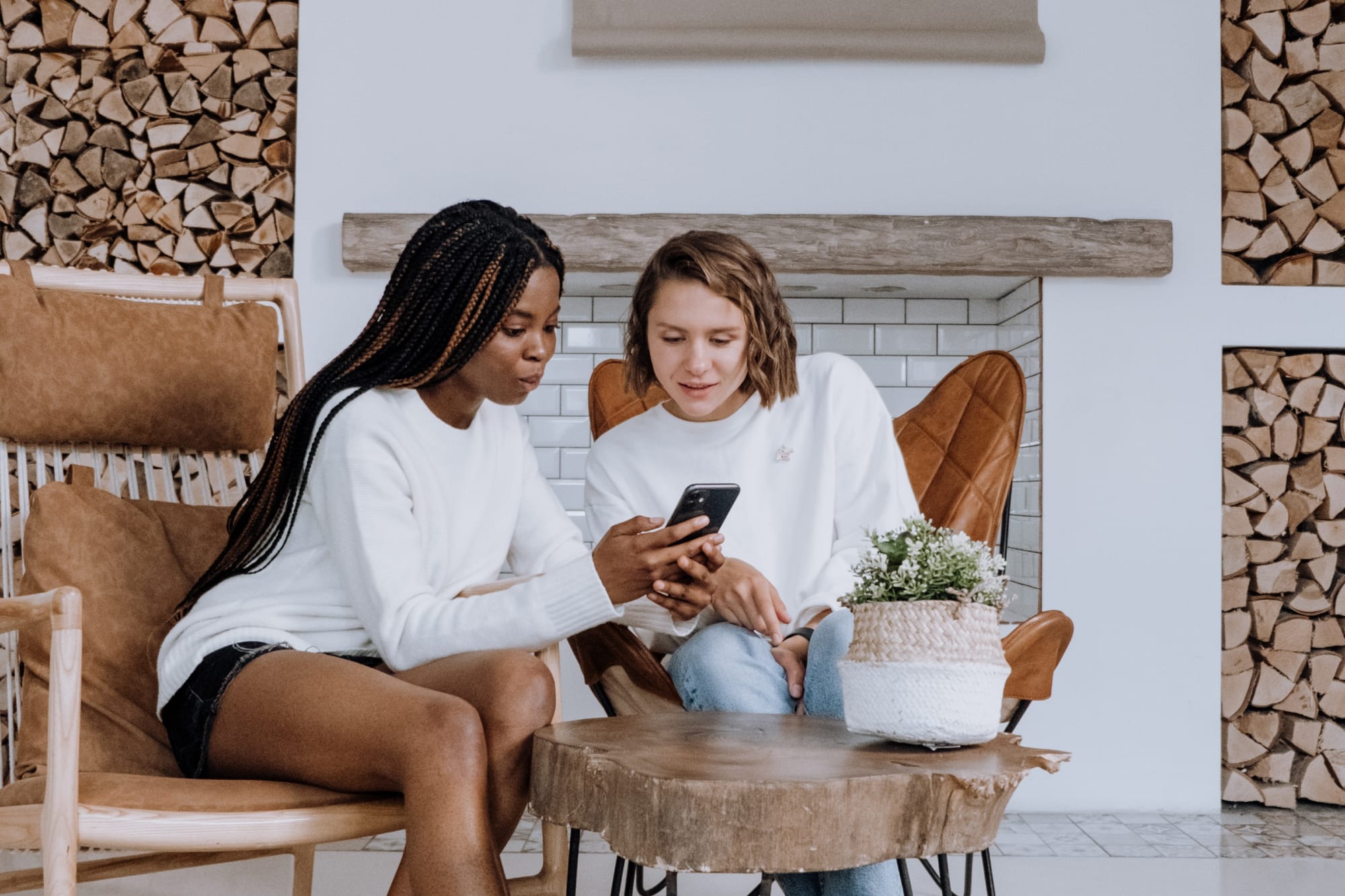 Two women sitting and looking at a smartphone together, with a rustic woodpile and fireplace in the background.